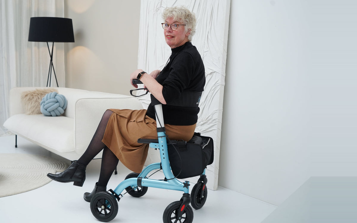 Vrouw zit op de lichtblauwe Safe Age rollator in een lichte woonkamer