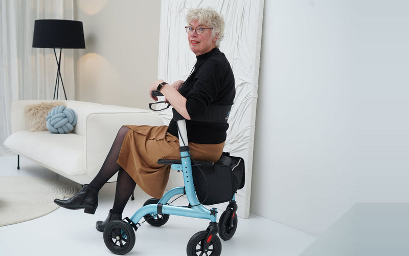 Vrouw zit op de lichtblauwe Safe Age rollator in een lichte woonkamer